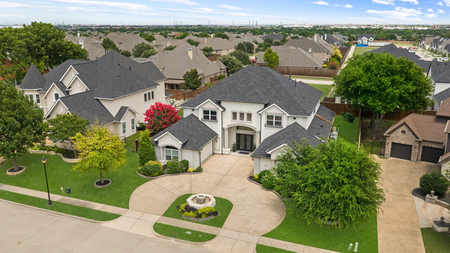 Drone photograph of a light stucco house, curved driveway and surrounding neighborhood
